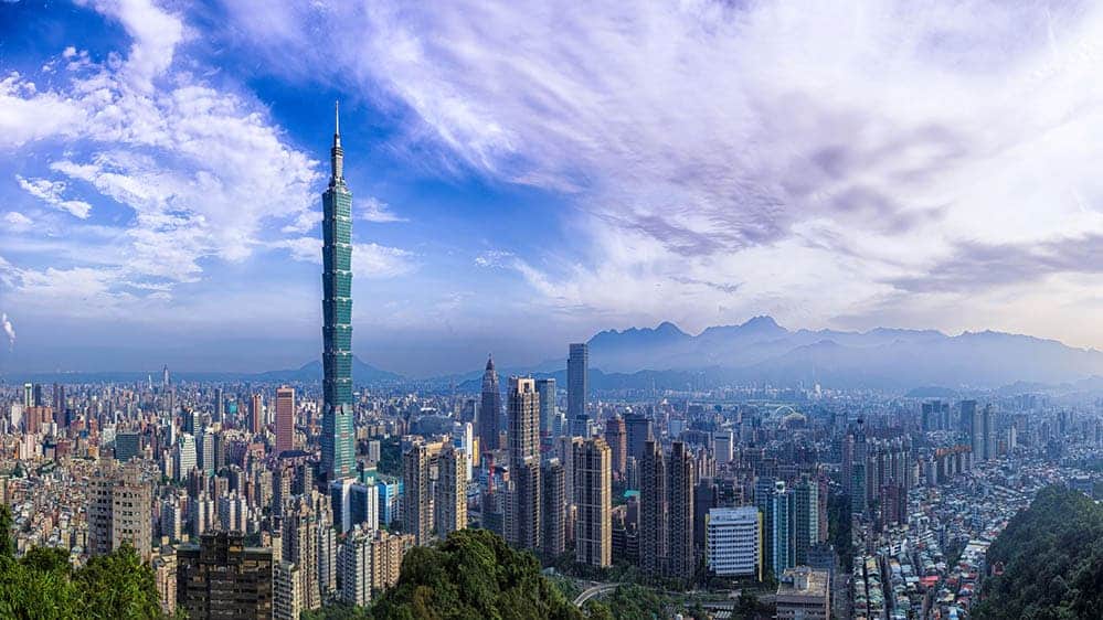 Panoramic view of Taipei City with Taipei 101 skyscraper and surrounding buildings, set against a backdrop of mountains and a partly cloudy sky, representing Taiwan as a top destination for international relocation.