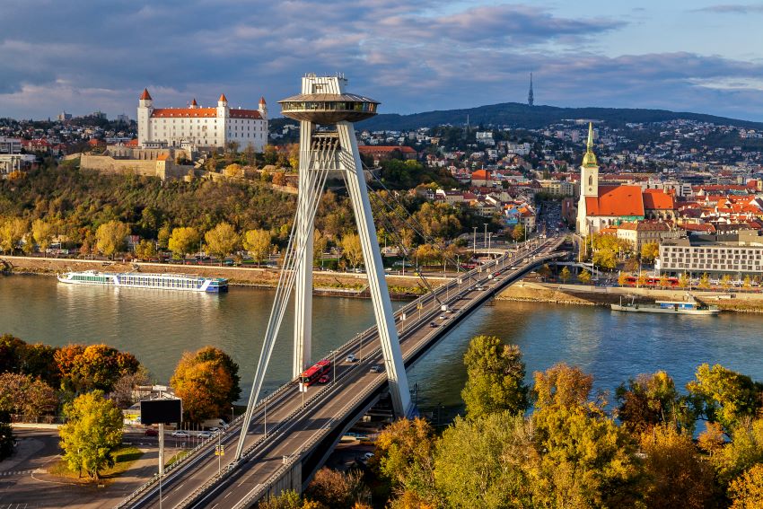 View of Bratislava, Slovakia featuring the iconic UFO Bridge crossing the Danube River with Bratislava Castle in the background amidst autumn-colored trees.