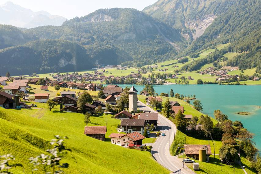 A scenic view of a Swiss village nestled by a lake, with lush green hills and mountains in the background, showcasing the natural beauty and tranquility of life in Switzerland.






