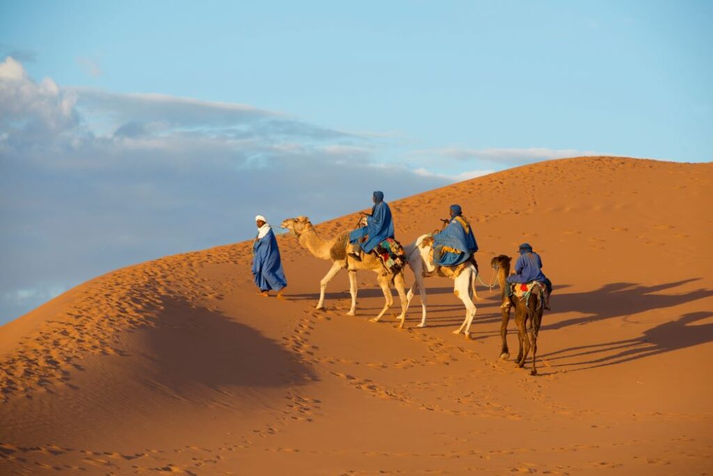 Three camel riders in blue robes traverse golden sand dunes, capturing the beauty and calm of a desert landscape, symbolizing the diversity and adventure of international relocation.