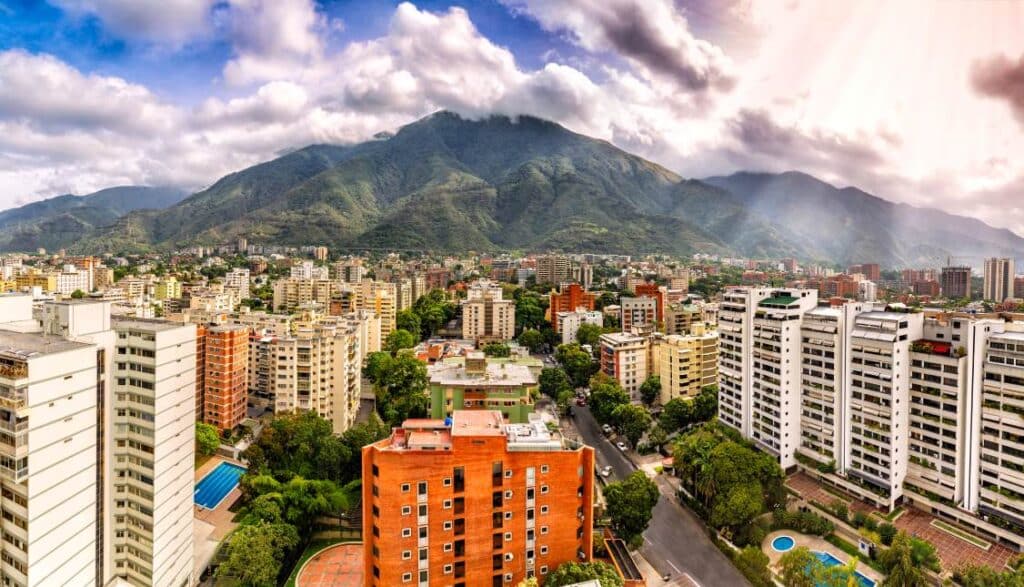 Panoramic view of Caracas, Venezuela, featuring tall buildings, tree-lined streets, and a backdrop of lush green mountains under a cloudy sky.