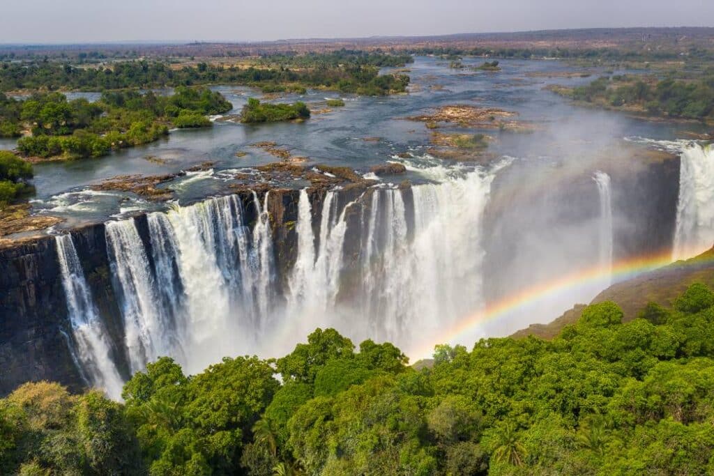 Panoramic view of Victoria Falls with water cascading down steep cliffs, surrounded by lush greenery and a rainbow forming in the mist.