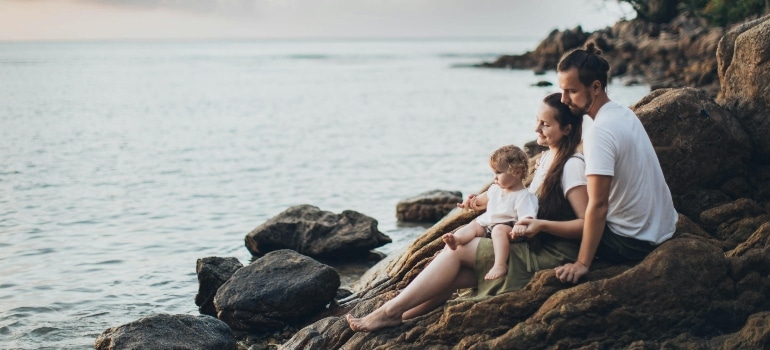 A couple, raising a family in Iceland, with their young child sits peacefully on coastal rocks, watching the sunset over the ocean while enjoying the quiet moments of togetherness.