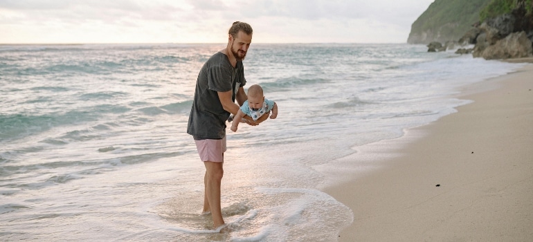 A father holds his baby above the shoreline, smiling as the waves gently touch their feet.


